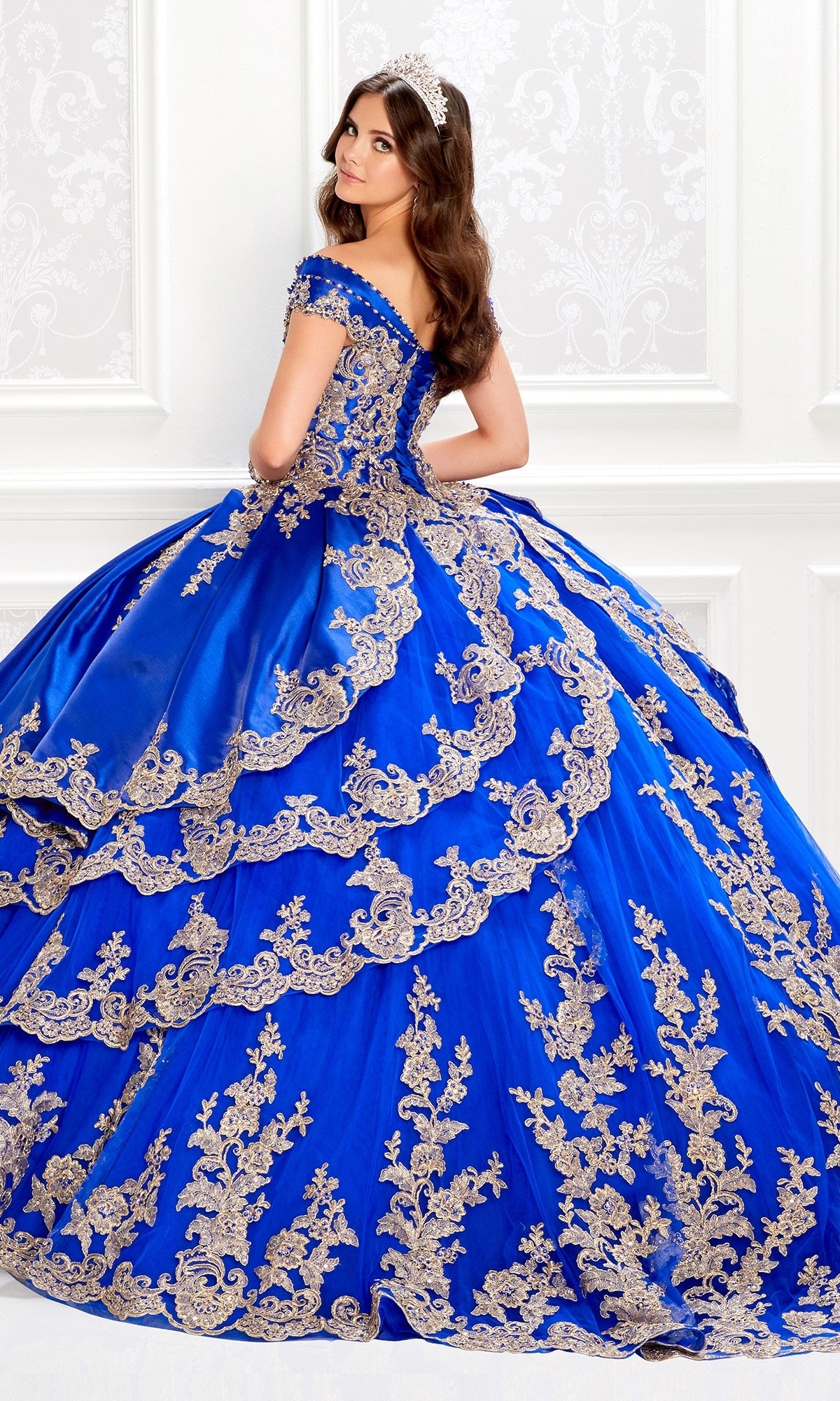 A woman in the Satin Quinceanera Dress: Princesa PR22029—a royal blue off-the-shoulder gown with gold embroidery—stands in an ornate white room, gazing over her shoulder.